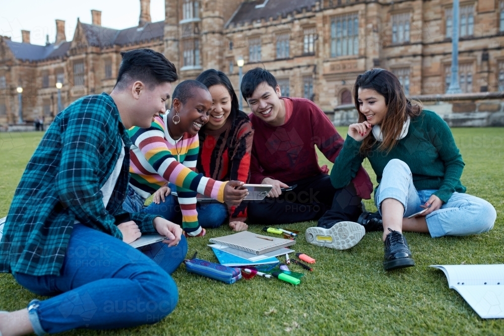 Group of young university students hanging out sitting on grass studying and using devices - Australian Stock Image