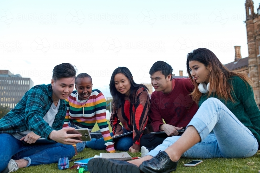 Group of young university students hanging out sitting on grass studying and using devices - Australian Stock Image