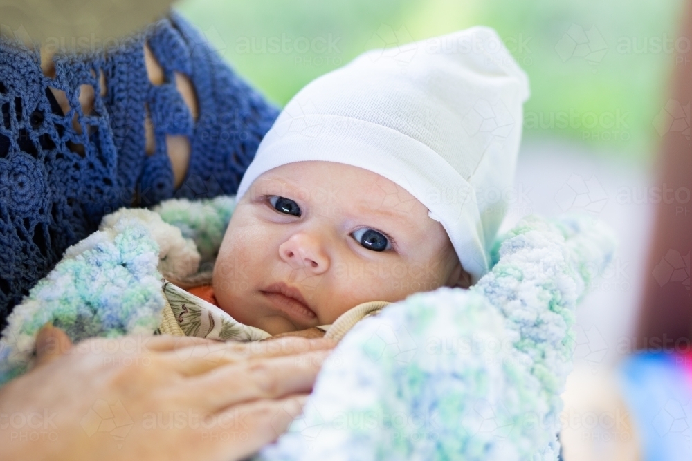 Grumpy baby face wrapped in blanket and beanie outside in cold held by grandma - Australian Stock Image