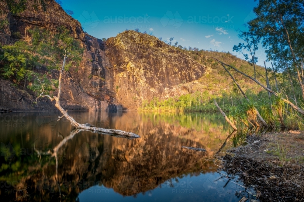 Beautiful golden reflections in a still lake with rocky escarpment in the background - Australian Stock Image