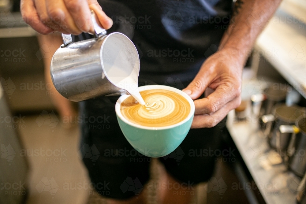 Half body shot of a barista pouring milk from a tin jar to the coffee in a green mug  making an art - Australian Stock Image