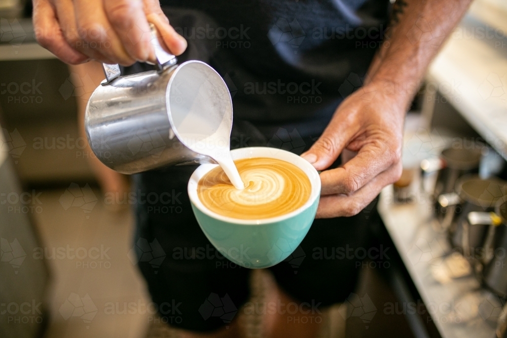 Half body shot of a barista pouring milk from a tin jar to the coffee in a green mug  making an art - Australian Stock Image