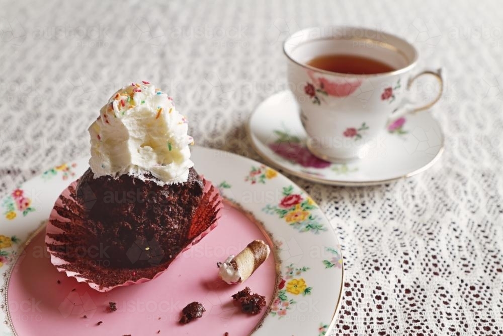 Half eaten chocolate cupcake with cup of tea in vintage crockery on lace tablecloth - Australian Stock Image