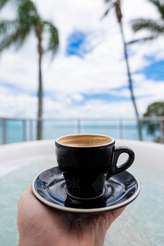 Hand holding an espresso cup with water and palm trees in background - Australian Stock Image
