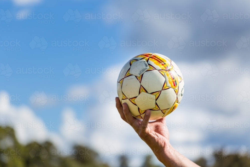Hand holding soccer ball up with cloudy blue sky background - Australian Stock Image