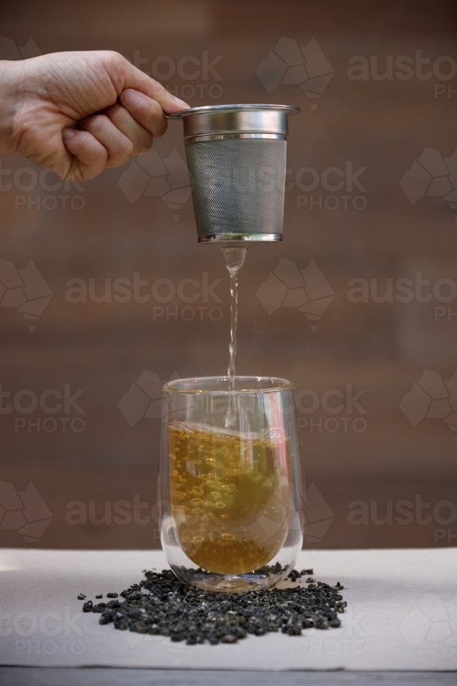 Hand pouring hot tea through strainer into glass cup - Australian Stock Image