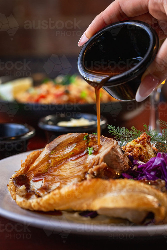 Hand pouring rich brown gravy from a small black jug onto a hearty pub‑style meal - Australian Stock Image