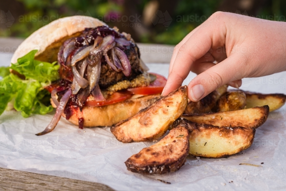 hand reaching for potato wedge, with hamburger in the background - Australian Stock Image