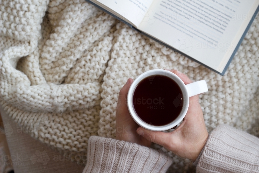 Hands holding coffee mug with woollen blanket and book - Australian Stock Image