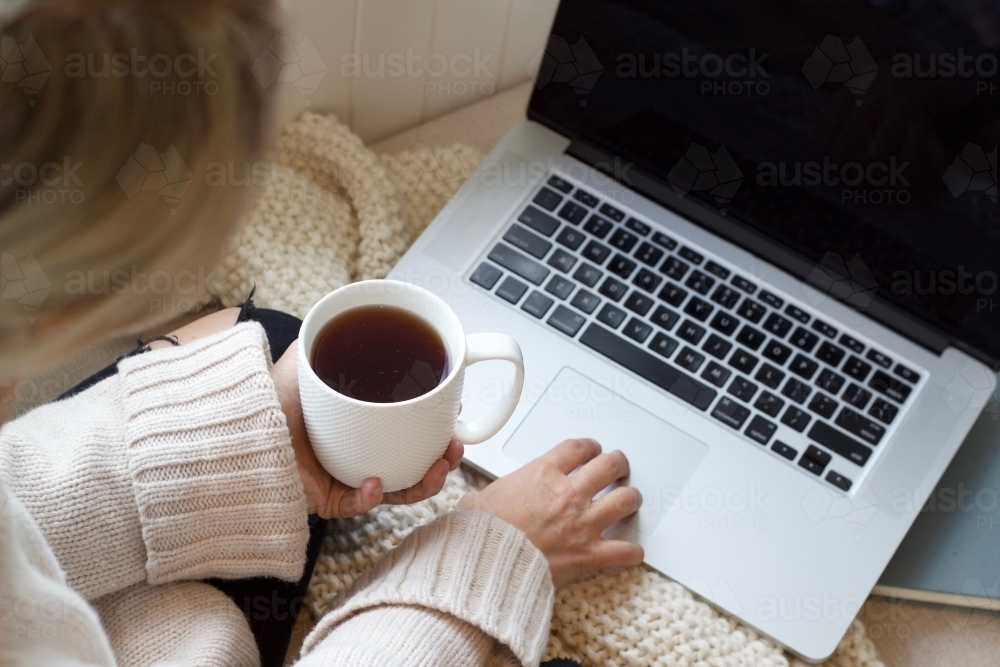 Hands holding coffee mug with woollen blanket and laptop computer and book - Australian Stock Image