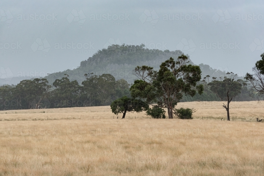 Hanging Rock with Summer Rain seen over dry paddock - Australian Stock Image