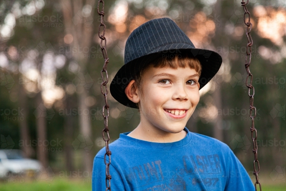 Happy 9yo kid in a hat playing on swing outside - Australian Stock Image