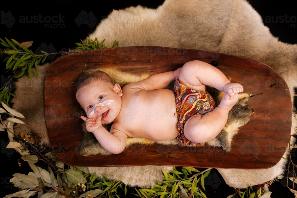 Happy Aboriginal baby boy lying on possum skin in coolamon cradle - Australian Stock Image