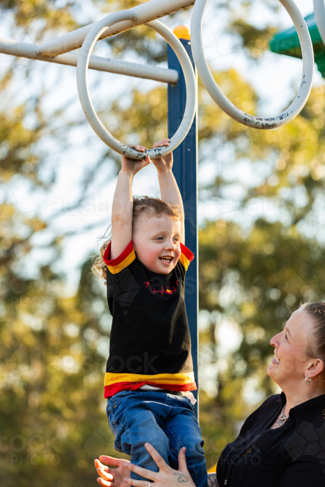 Happy Aboriginal toddler boy playing on monkey bar rings at park with mum - Australian Stock Image
