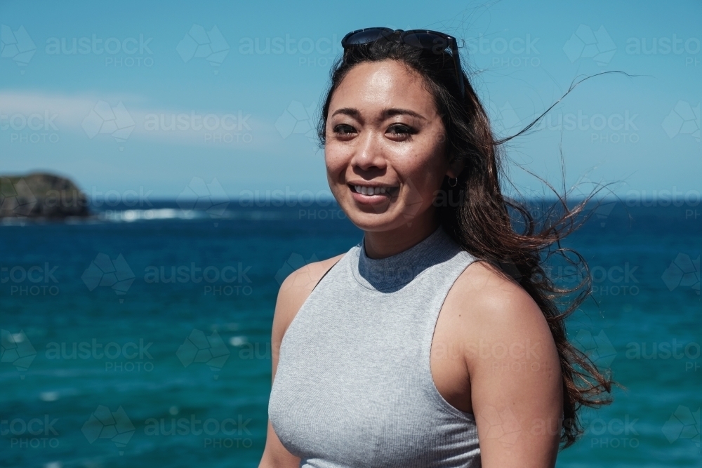 Happy Asian young adult woman with blue ocean background on summer day - Australian Stock Image