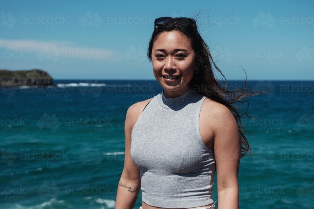 Happy Asian young adult woman with blue ocean background on summer day - Australian Stock Image