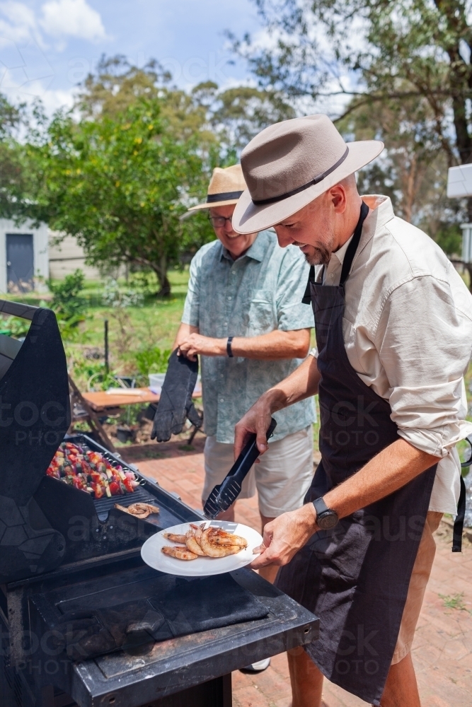 happy Aussie men cooking lunch food on bbq in backyard of country home - Australian Stock Image