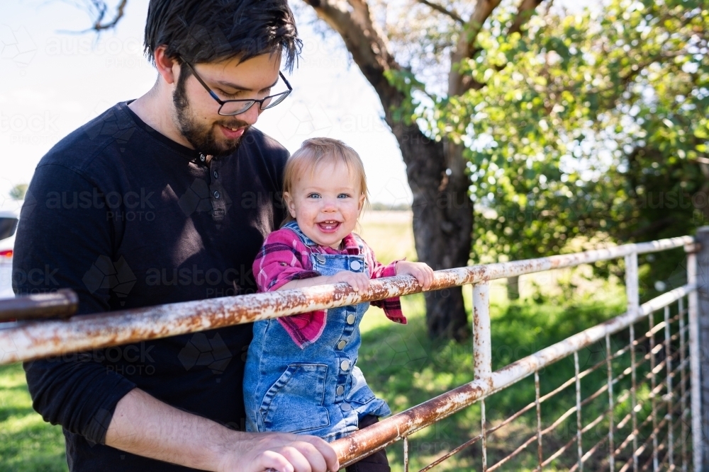 Happy baby country kid swinging on farm gate with dad - Australian Stock Image