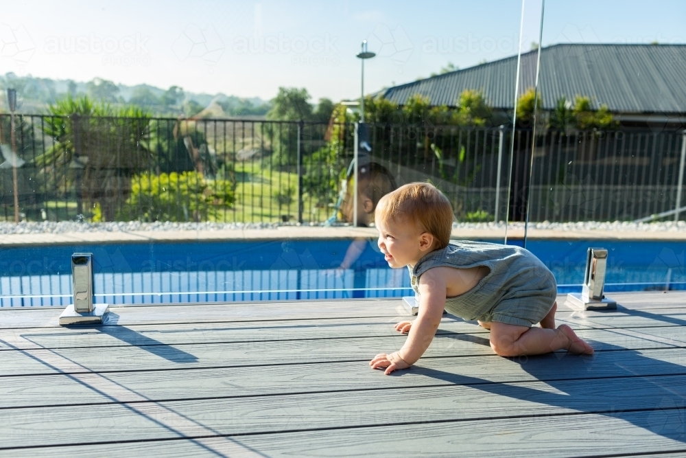 Happy baby crawling on deck beside glass pool fence in summer - Australian Stock Image