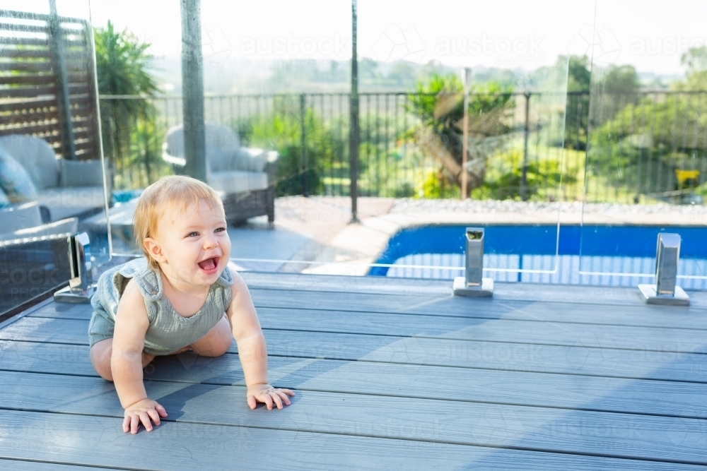happy baby crawling on deck beside inground pool in backyard - Australian Stock Image