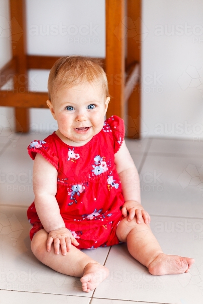 Happy baby girl in red Christmassy outfit sitting on kitchen floor - Australian Stock Image
