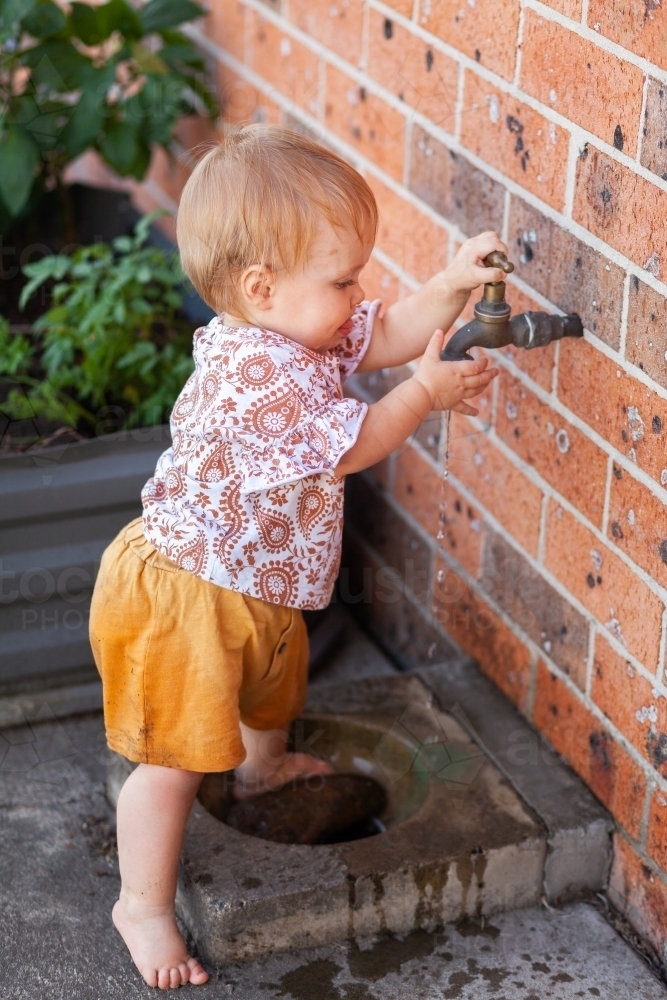 Happy baby girl outside in backyard playing in tap water in summer - Australian Stock Image