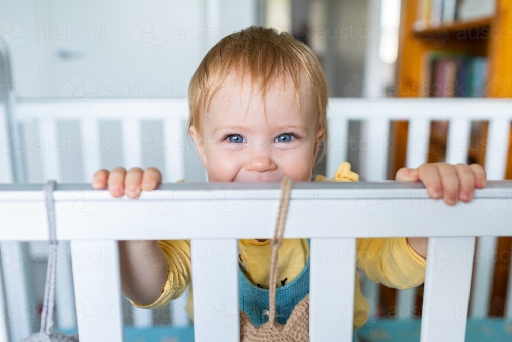 Image of happy baby girl peering over cot bars in bedroom Austockphoto