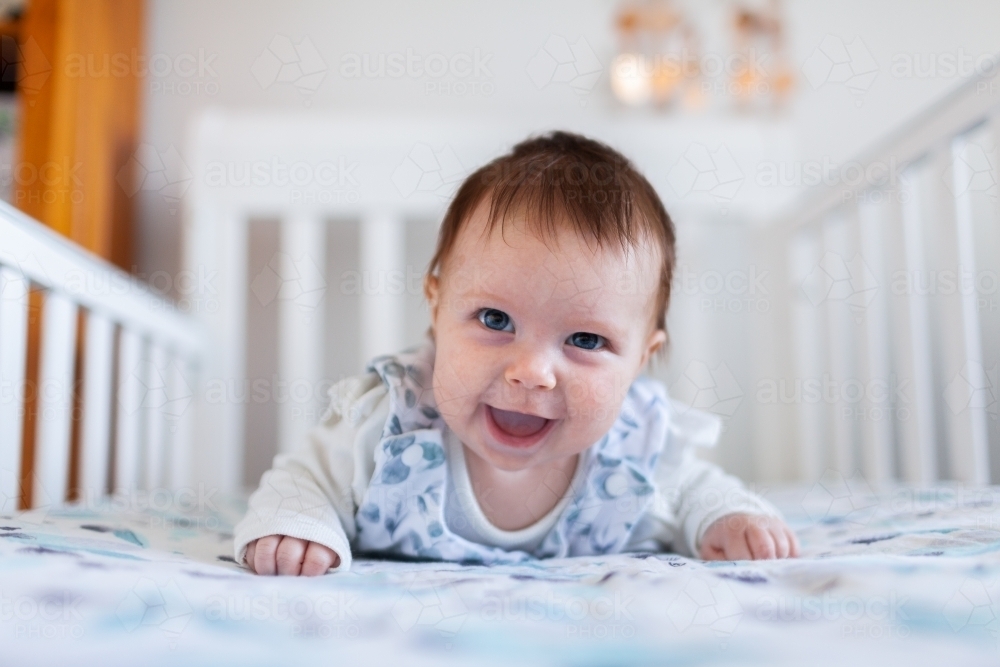 Happy baby grinning doing tummy time in crib in bedroom - Australian Stock Image