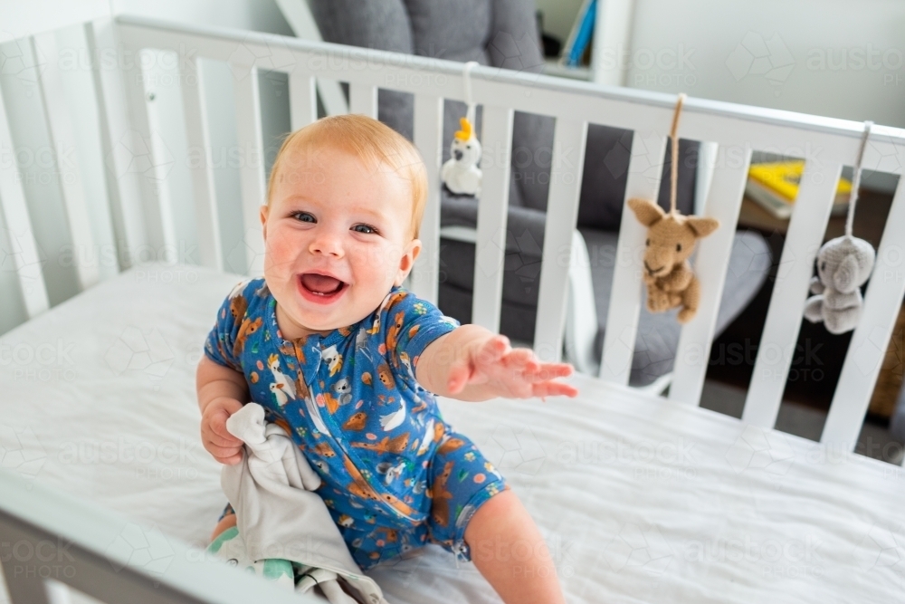 Happy baby in white cot with blanky toy reaching out - Australian Stock Image