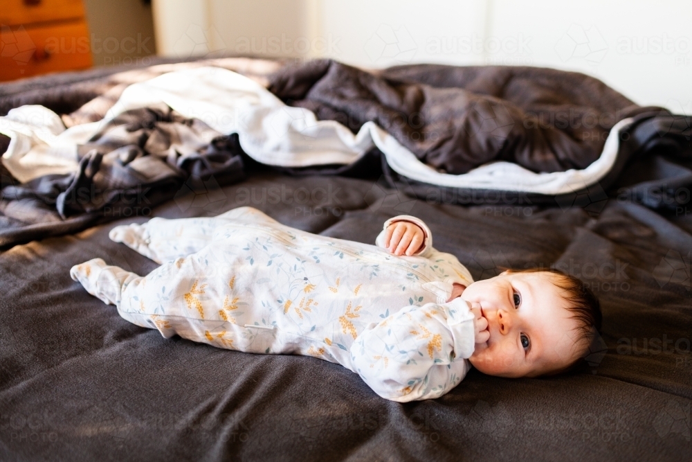 Happy baby lying on parents bed sucking fingers and smiling - Australian Stock Image