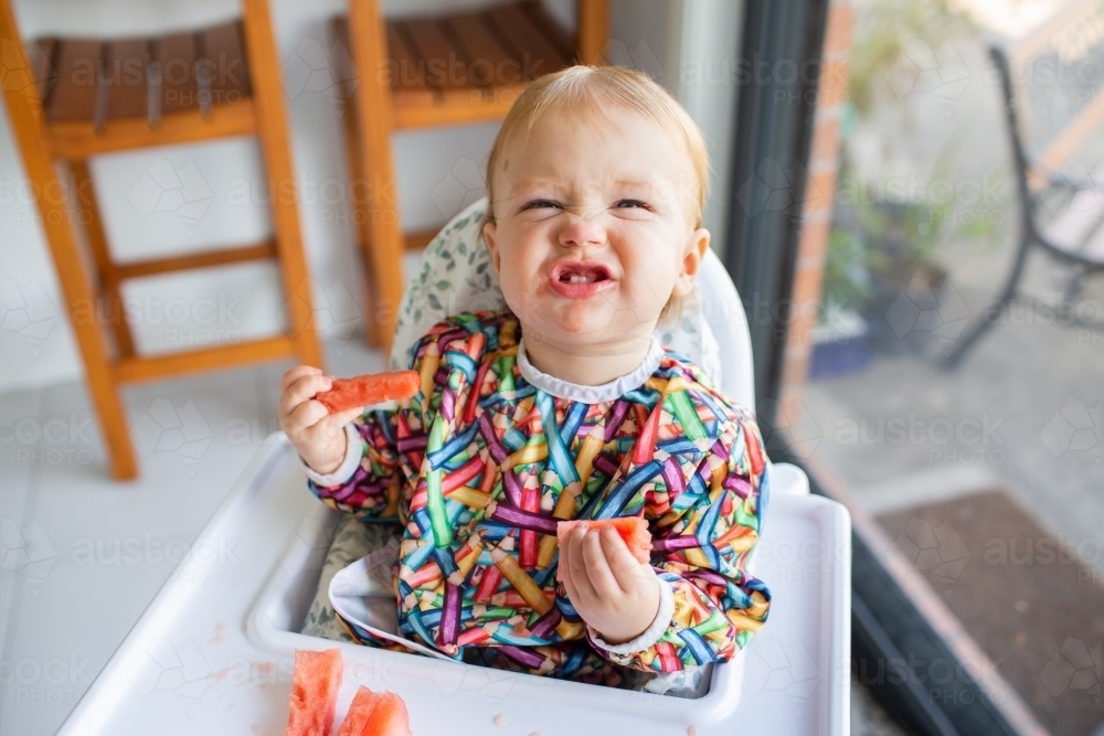 Image of Happy baby pulling silly face eating watermelon for a snack