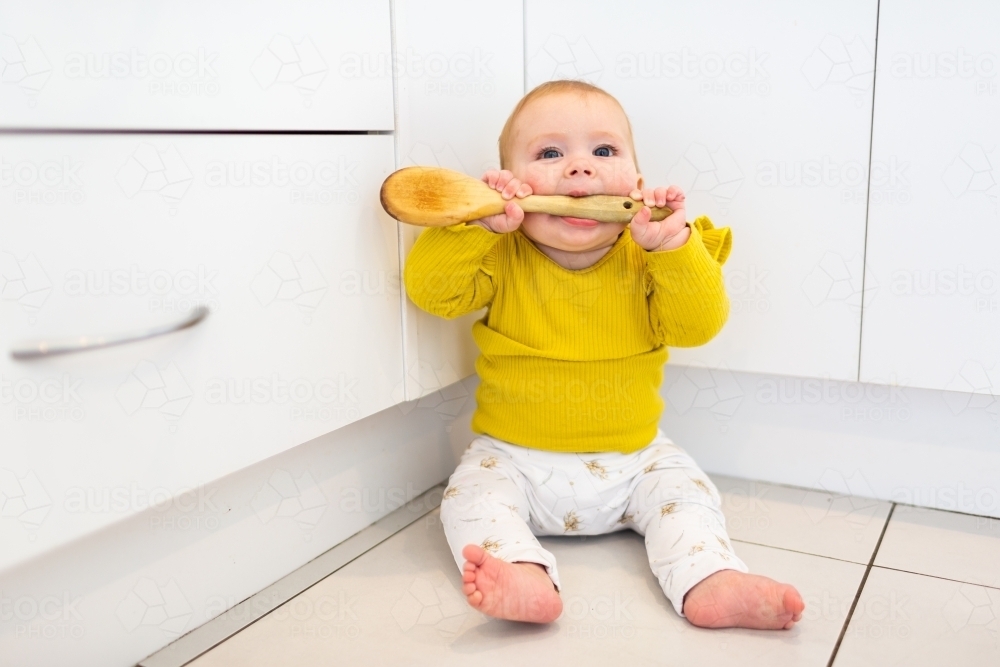 happy baby sitting in corner of kitchen playing with wooden spoon - Australian Stock Image