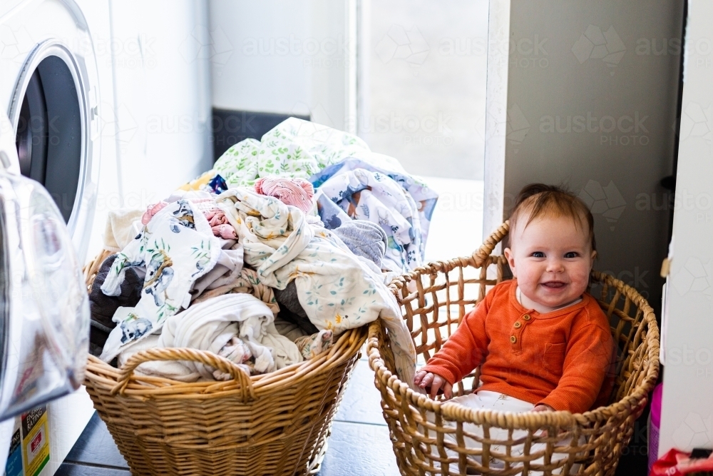 Happy baby sitting in laundry basket helping get wet clothes out of washing machine - Australian Stock Image