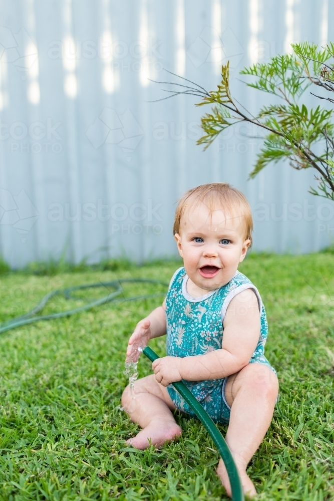 happy baby sitting on lawn in backyard with garden hose - Australian Stock Image