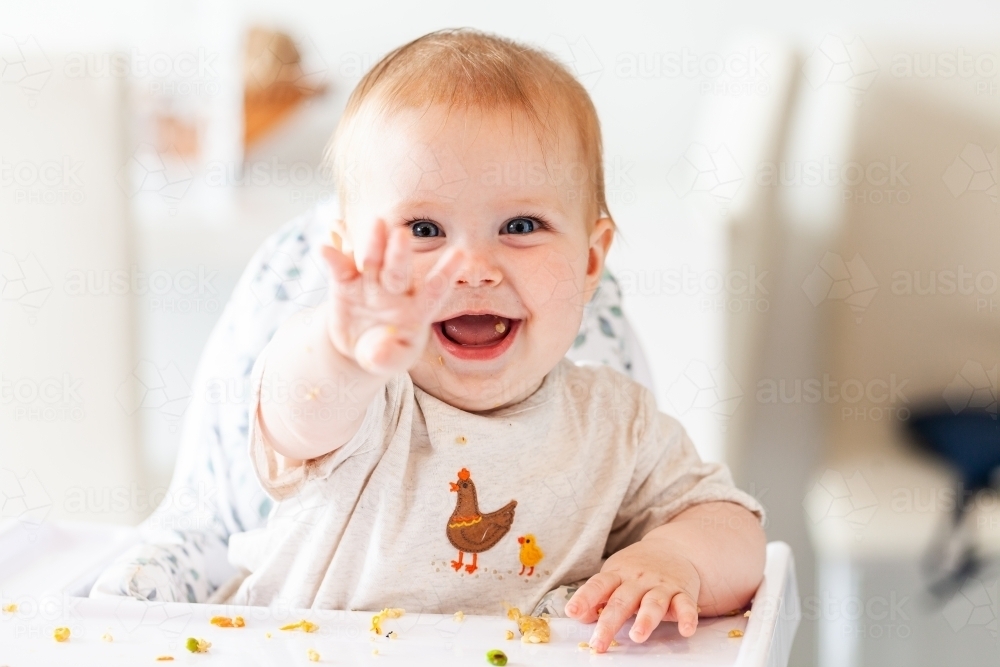 Happy baby smiling in high chair pointing with scraps of food - Australian Stock Image
