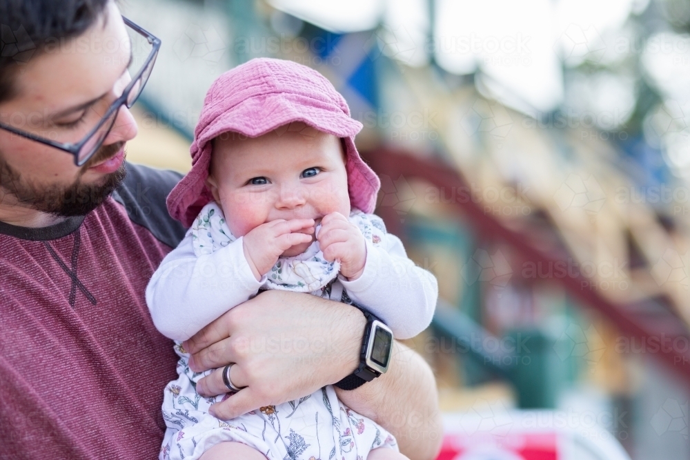 Image of Happy baby wearing hat outside in dads arms with grandstand