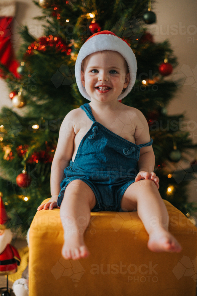 Happy blonde toddler sits on a yellow stool with a joyous grin by Christmas decorations - Australian Stock Image