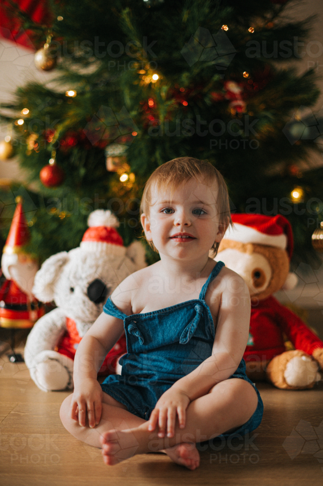 happy blonde toddler smiles by a teddy bear and Christmas tree - Australian Stock Image
