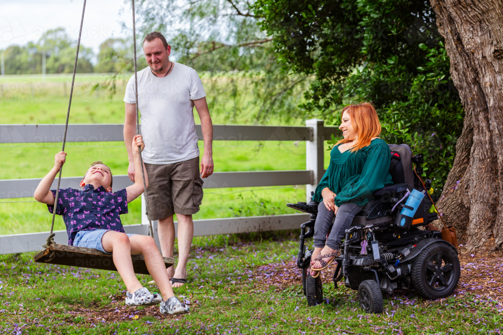 Happy boy on swing while dad pushes and mum watches from motorised wheelchair - Australian Stock Image