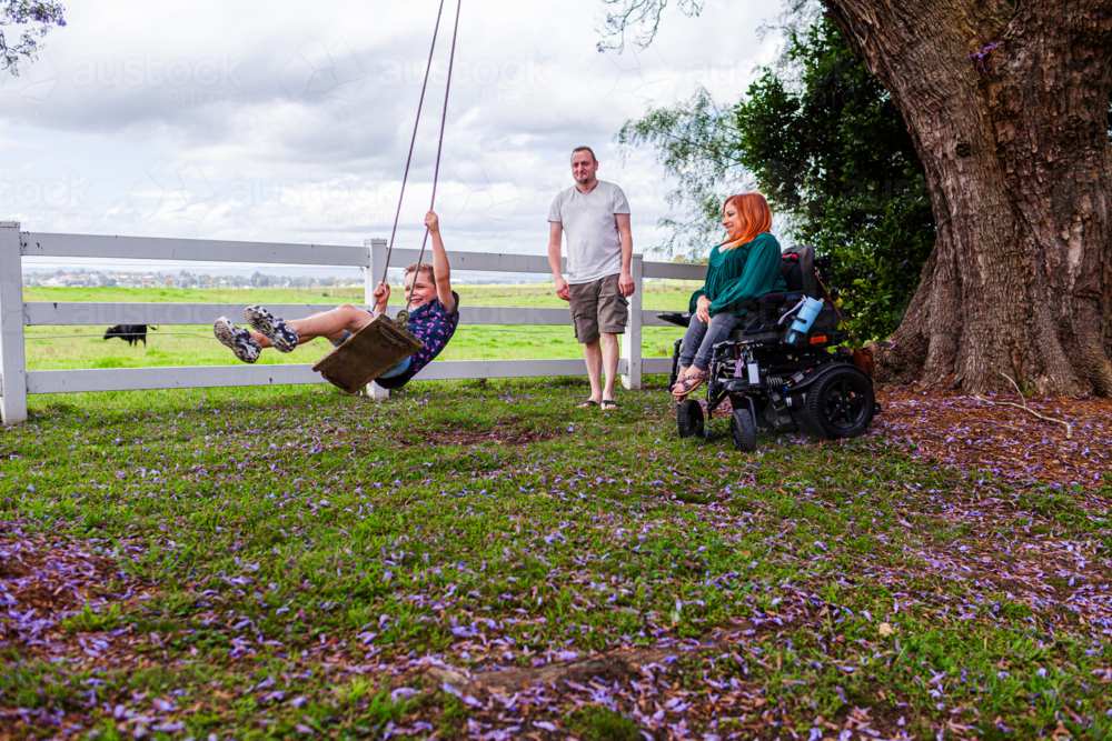 Happy boy on swing while dad pushes and mum watches from motorised wheelchair - Australian Stock Image