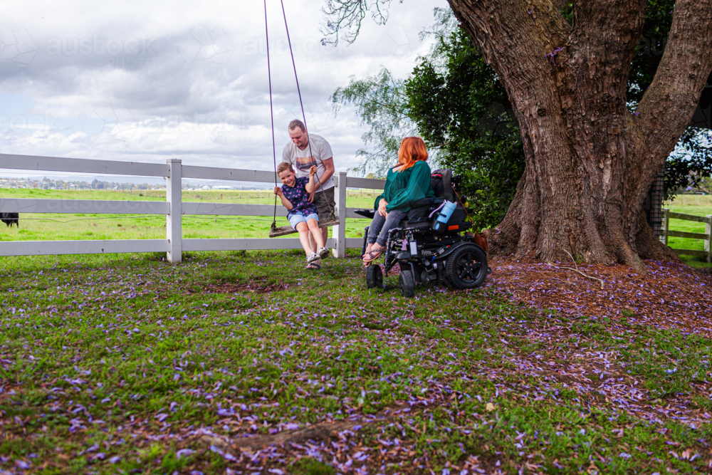 Happy boy on swing while dad pushes and mum watches from motorised wheelchair - Australian Stock Image