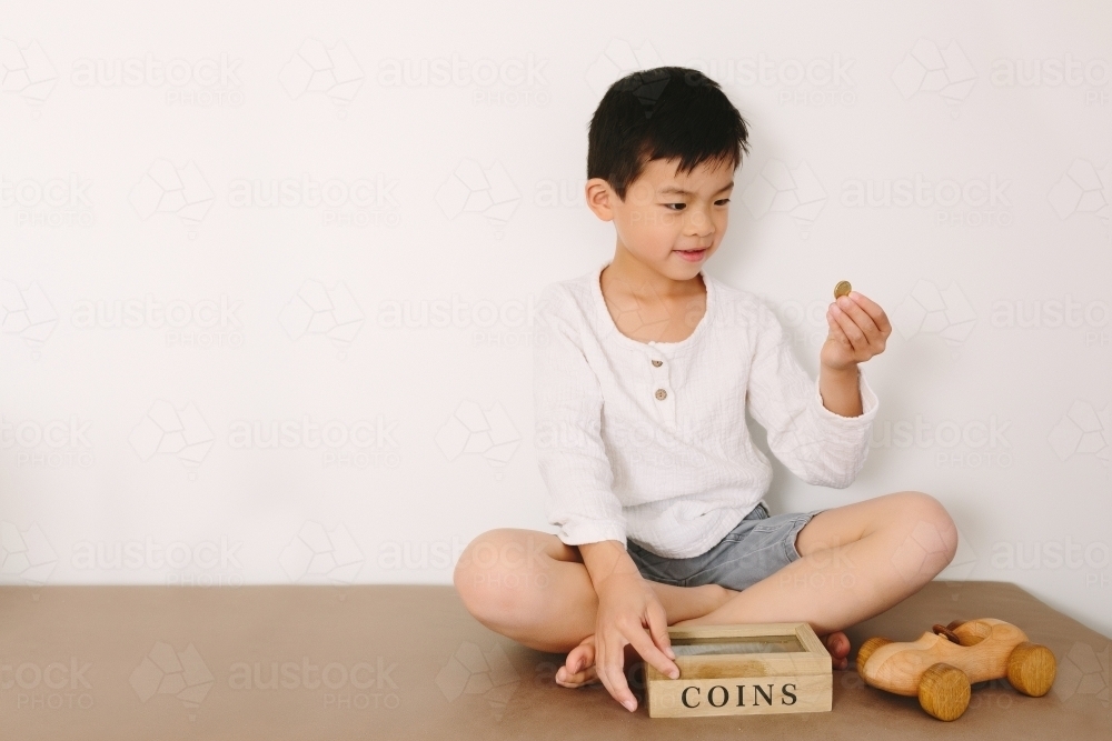 Happy boy sitting down putting money into his coin box - Australian Stock Image