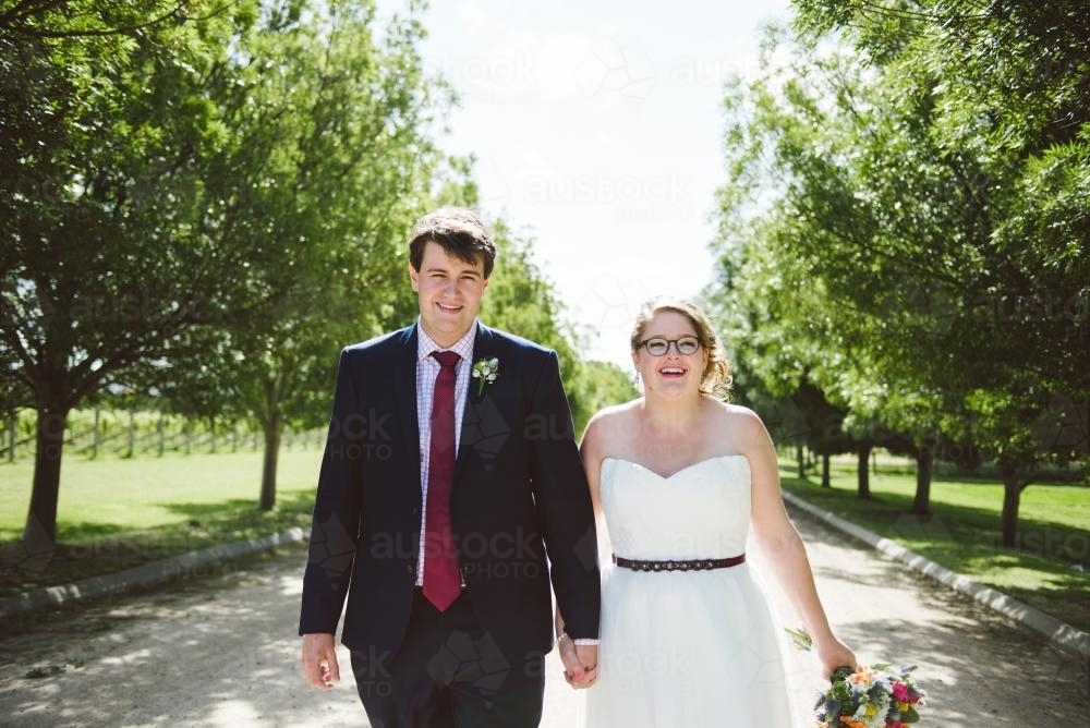 Happy Bride and Groom walking along road through green trees - Australian Stock Image