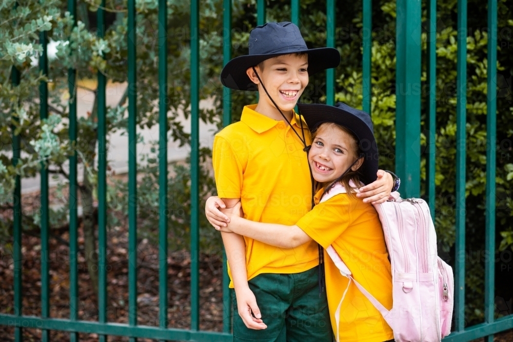 Happy brother and sister laugh and hug beside school fence outside - Australian Stock Image