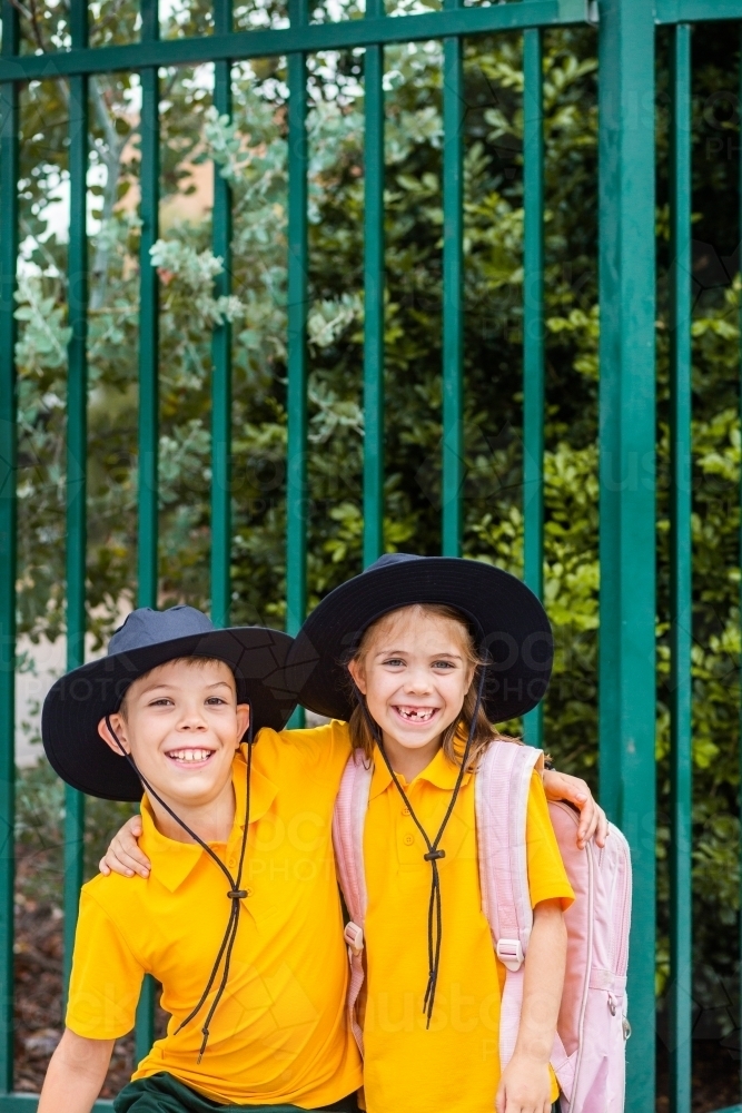 Happy brother and sister laugh and hug beside school fence outside - Australian Stock Image