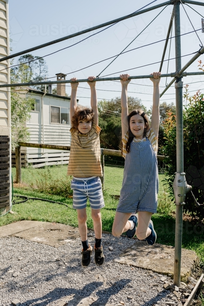 Happy brother & sister hanging from a Hills Hoist clothes line in an Australian country backyard - Australian Stock Image