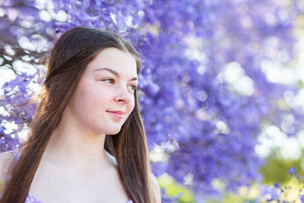 Happy brunette teenage girl standing among flowering branches of purple jacaranda tree - Australian Stock Image