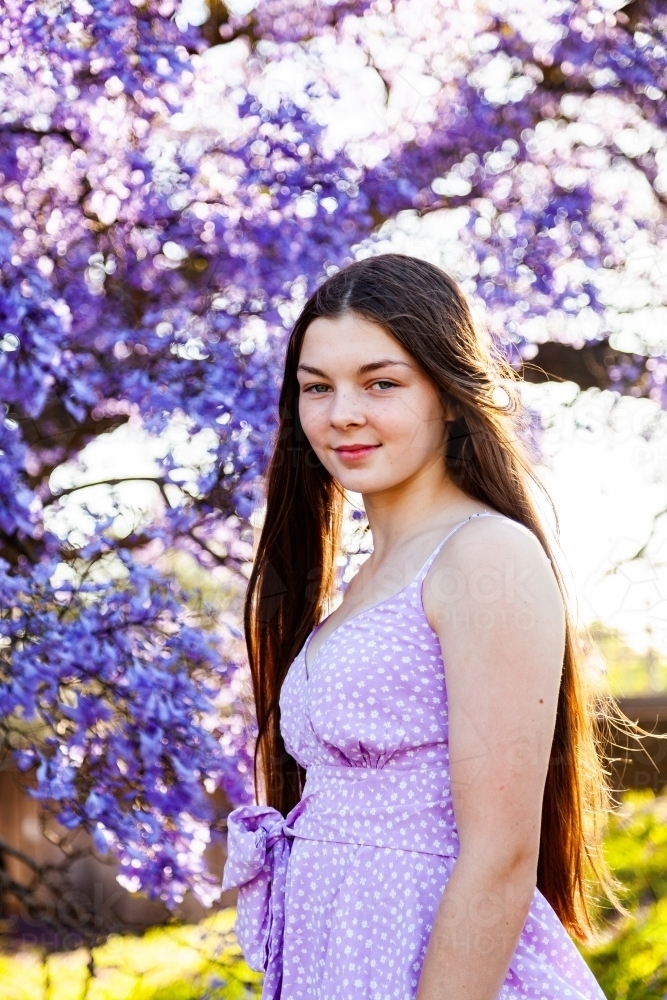 Happy brunette teenage girl standing among flowering branches of purple jacaranda tree - Australian Stock Image