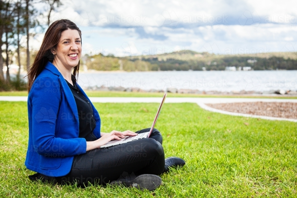 Portrait of happy business woman smiling  with laptop in park sitting on grass - Australian Stock Image