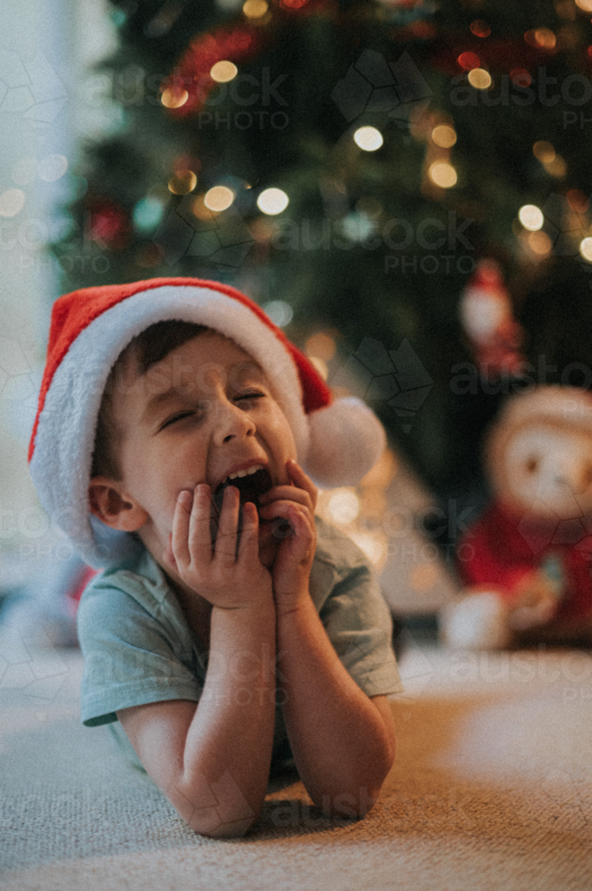 happy buy lying by christmas tree - childhood joy during Christmas - Australian Stock Image